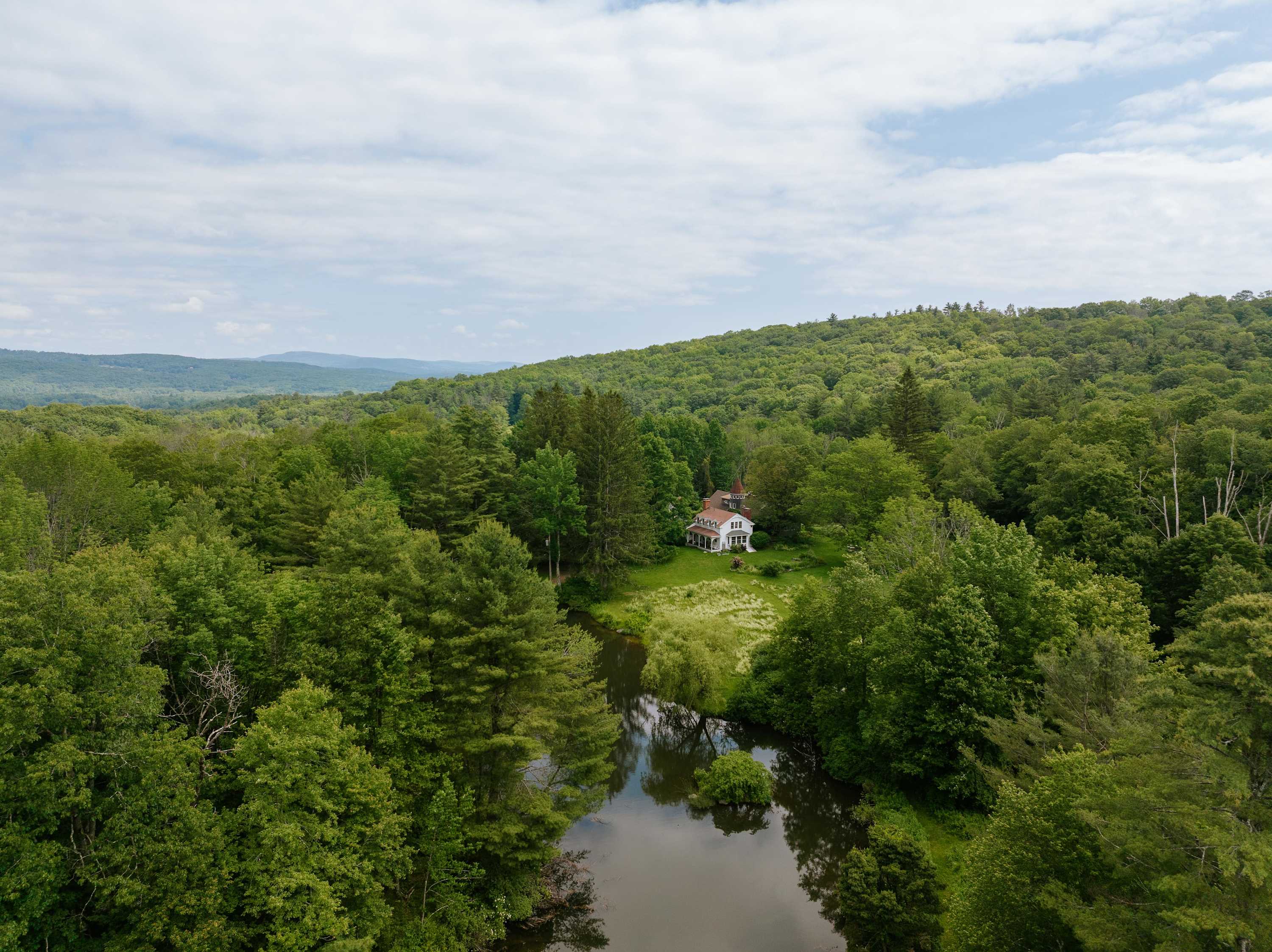 Panoramic aerial view showcasing the white house nestled within a sprawling, dense green forest, with a calm river or pond reflecting the sky in the foreground, highlighting the secluded natural setting.