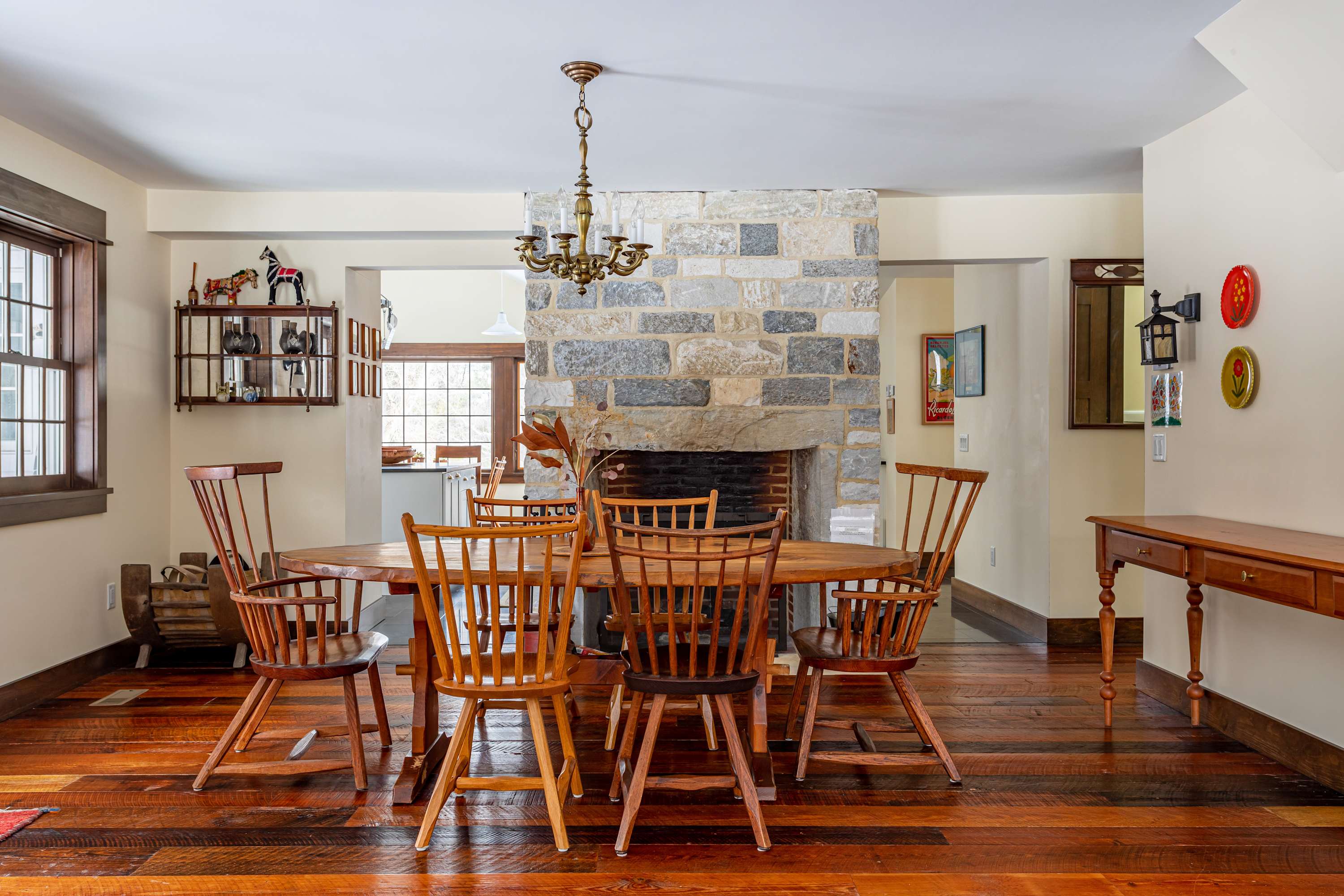 Rustic dining room with polished hardwood floors, a large oval wooden dining table surrounded by Windsor chairs, and a prominent stone fireplace as a central feature.