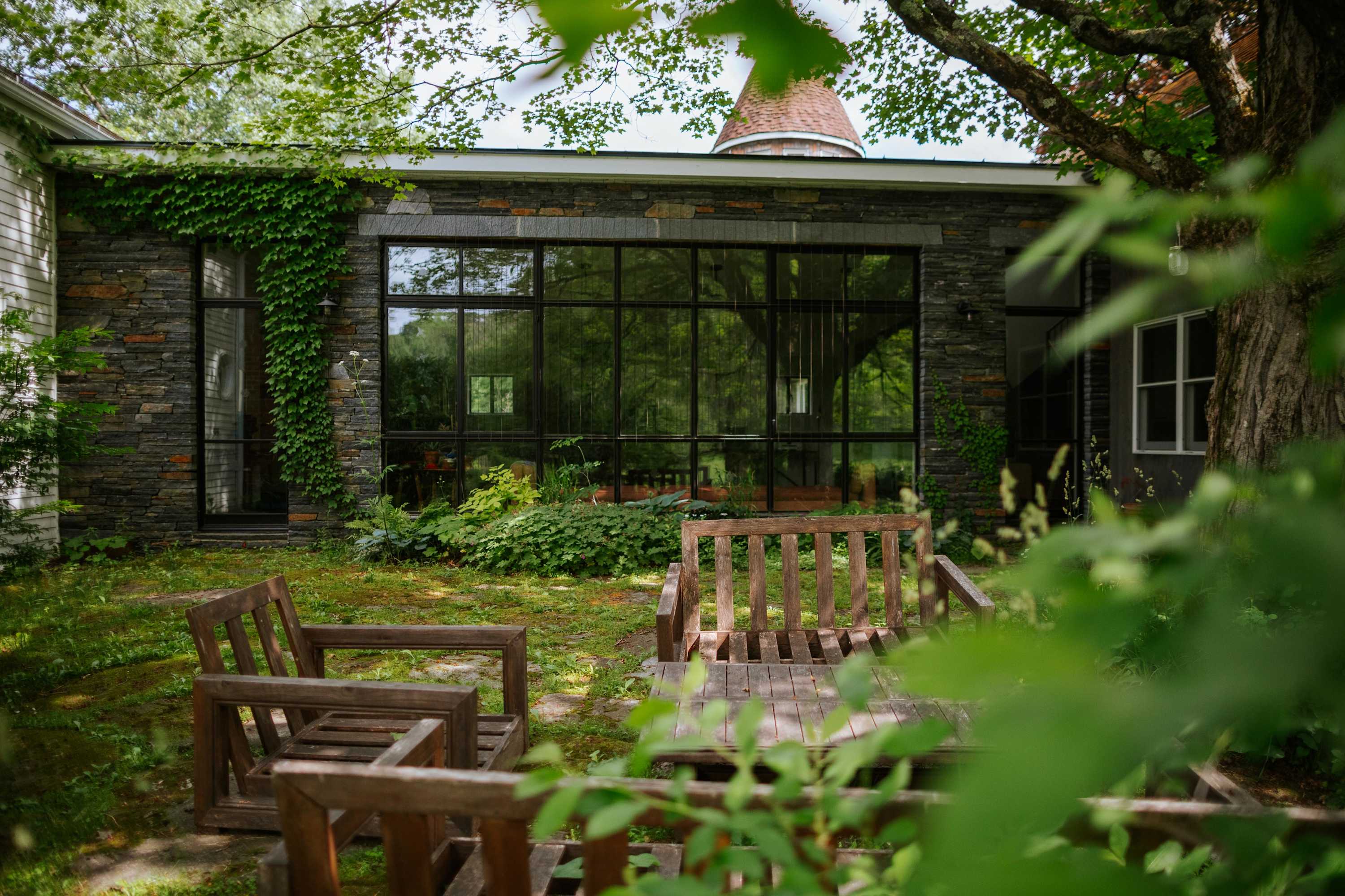 A stone patio area with large windows reflecting lush greenery is surrounded by wooden chairs and a wooden bench, set amidst a grassy yard with mature trees. The building features a mix of stone and wood siding.