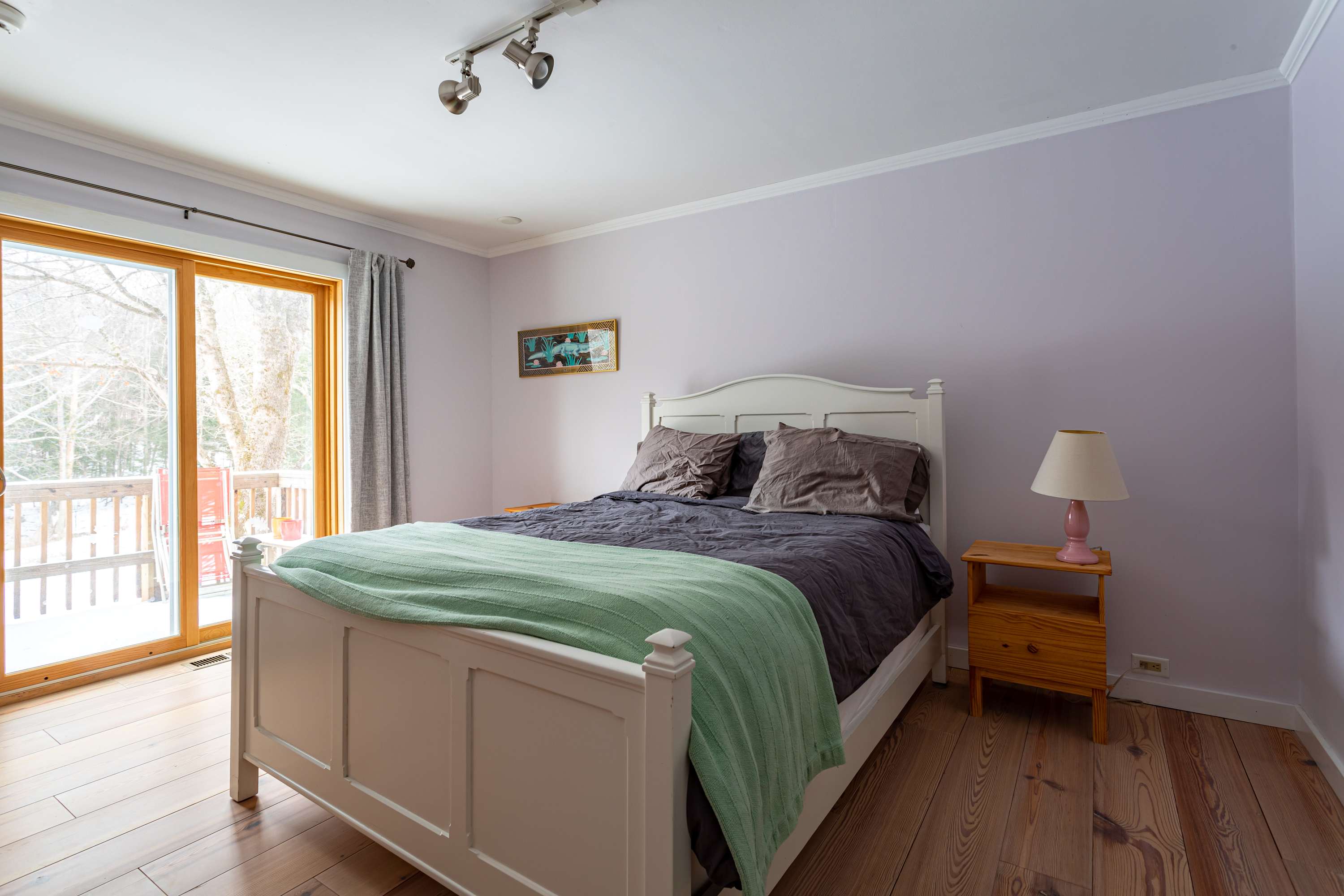 A serene bedroom with light lavender walls and hardwood floors features a white wooden bed frame with a dark gray duvet and a light green throw, flanked by a wooden nightstand with a pink lamp. Large glass doors lead to a snowy outdoor deck.