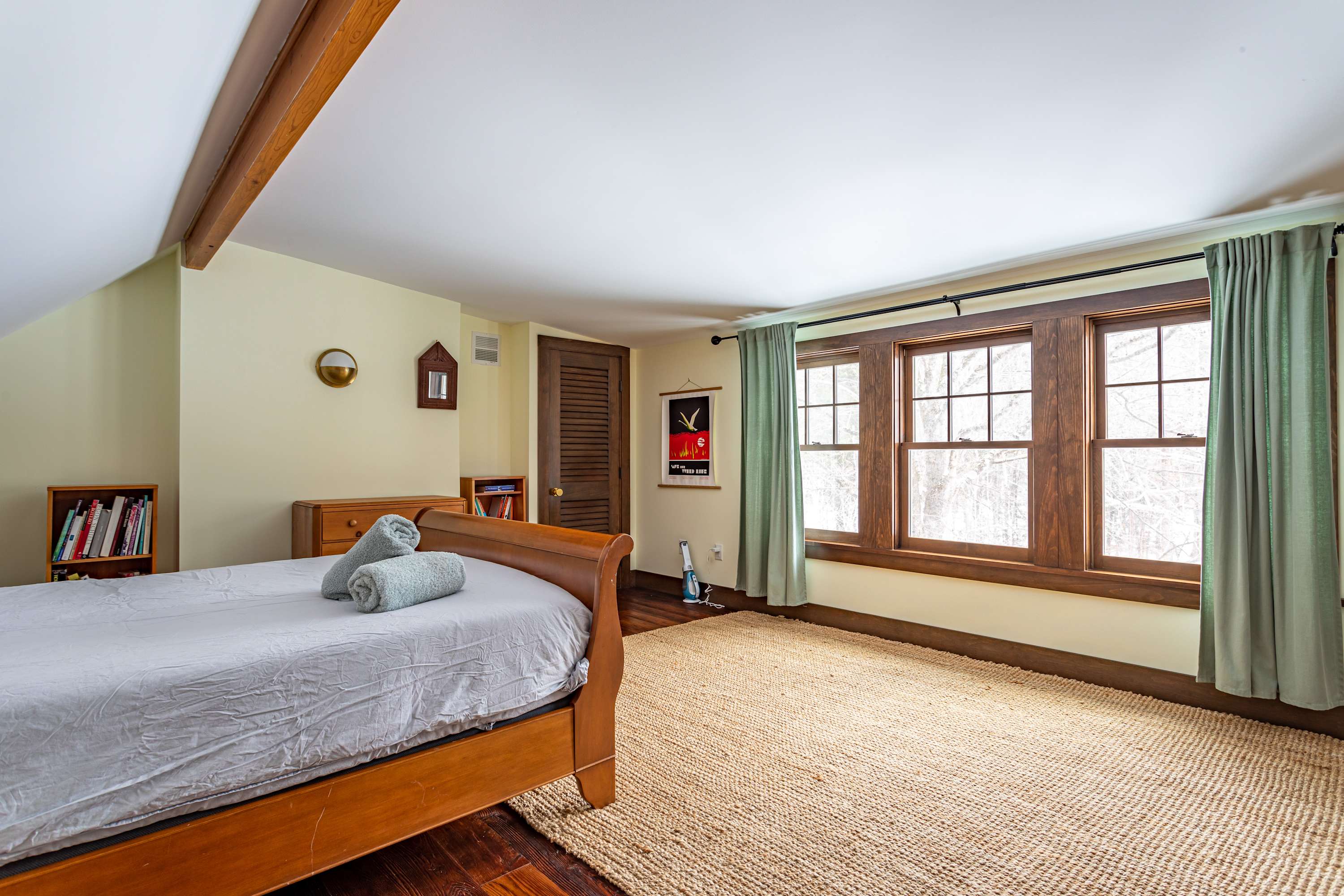 Attic bedroom featuring a wooden sleigh bed, dark hardwood floors, and light yellow walls. Three large windows with dark wood trim and light green curtains overlook a snowy landscape.