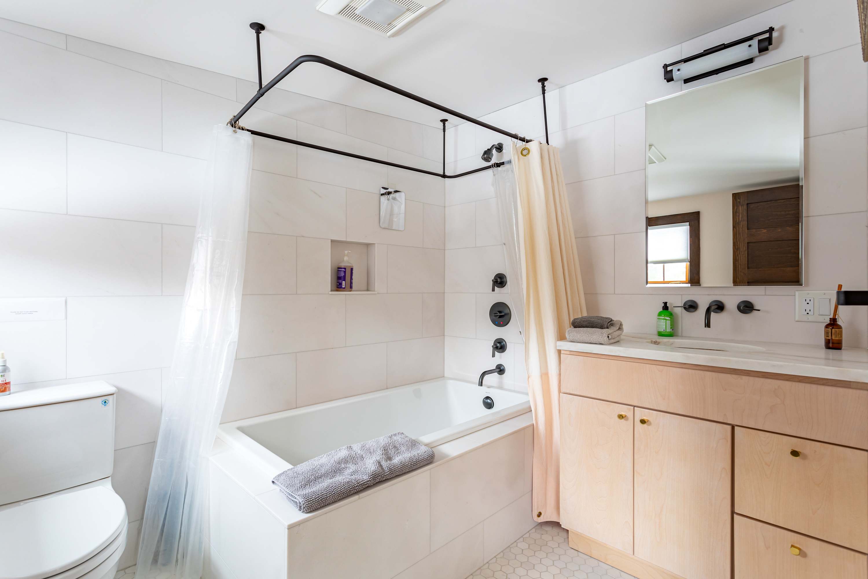 Modern bathroom with white subway tiles, a deep soaking tub with a black shower frame, and a light wood vanity with a white countertop and black fixtures. Hexagonal floor tiles are visible.