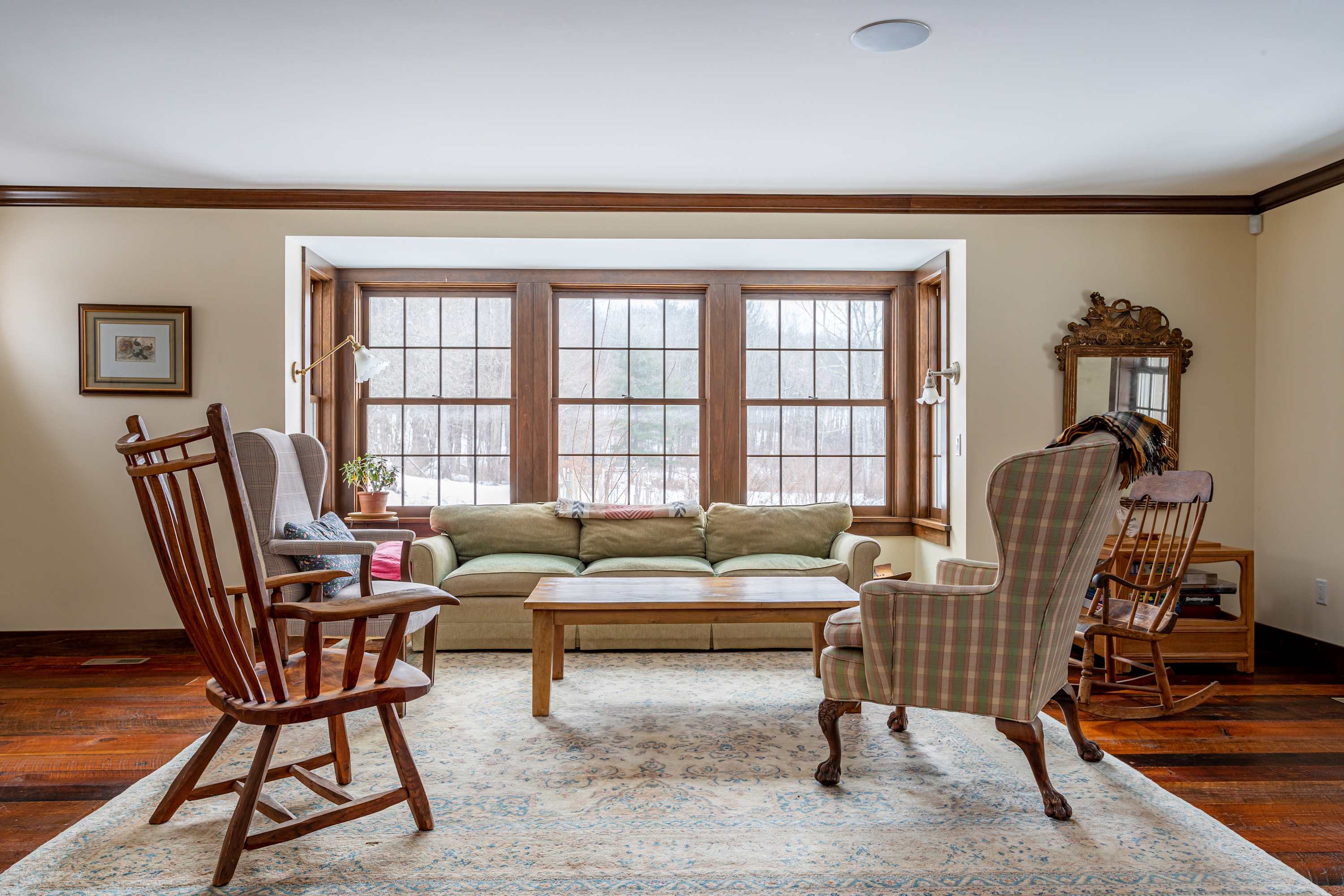 Bright living room with dark hardwood floors, a large patterned area rug, a light green sofa, and a wooden coffee table, facing a bay window with views of a snowy landscape.