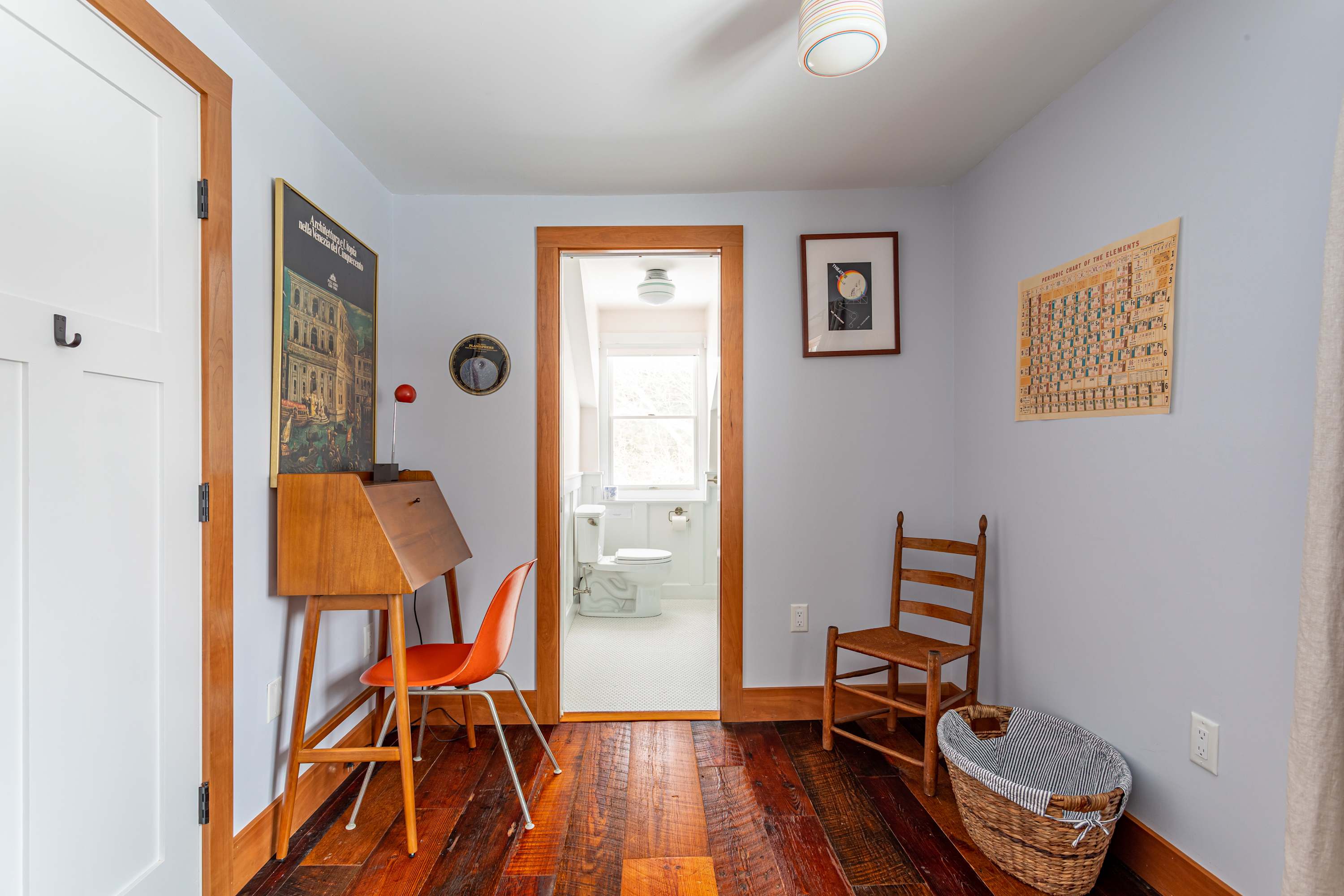 View of a small bedroom with warm hardwood floors, a wooden desk with an orange chair, and an open doorway leading to an en-suite bathroom.