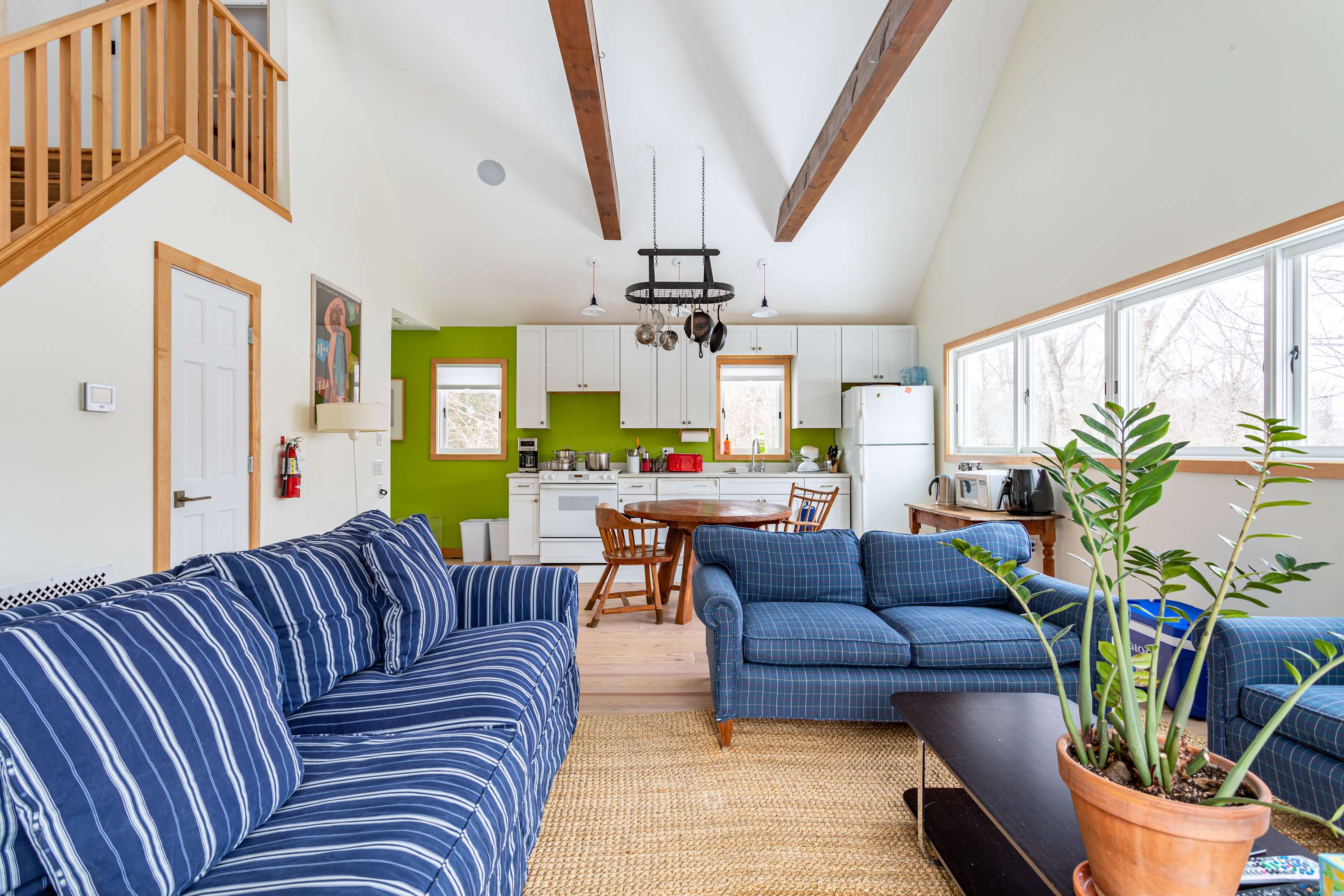 Open-concept living area with high vaulted ceilings, exposed wooden beams, and light hardwood floors. Two blue striped sofas face a dark coffee table, with a kitchen visible in the background.