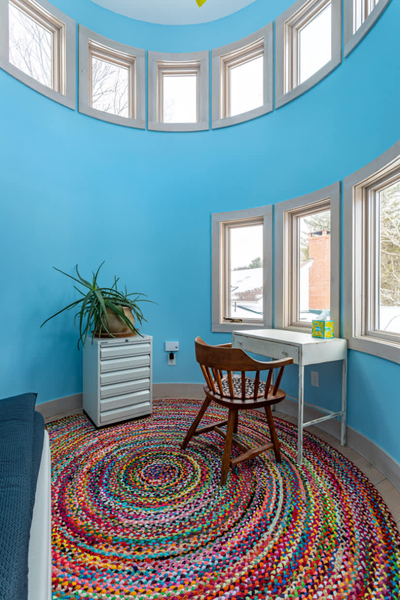 A unique circular office nook with bright blue walls and multiple small windows features a small white desk, a wooden chair, and a colorful circular braided rug. A plant sits on a metal cabinet.