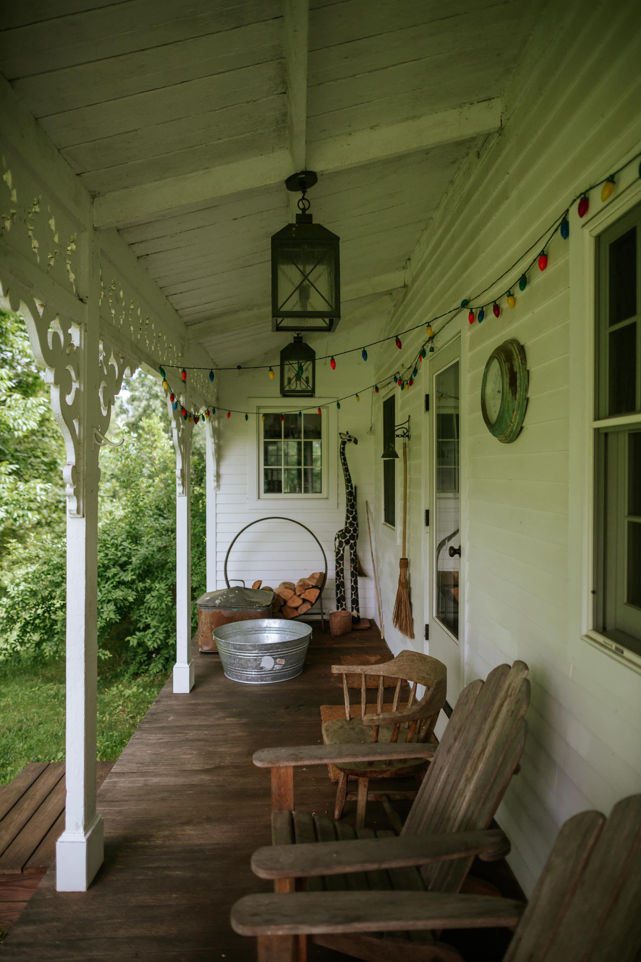A charming white wooden porch features two Adirondack chairs, a galvanized tub with firewood, and a tall giraffe sculpture, all under a string of colorful lights and a hanging lantern. The porch overlooks lush green foliage.