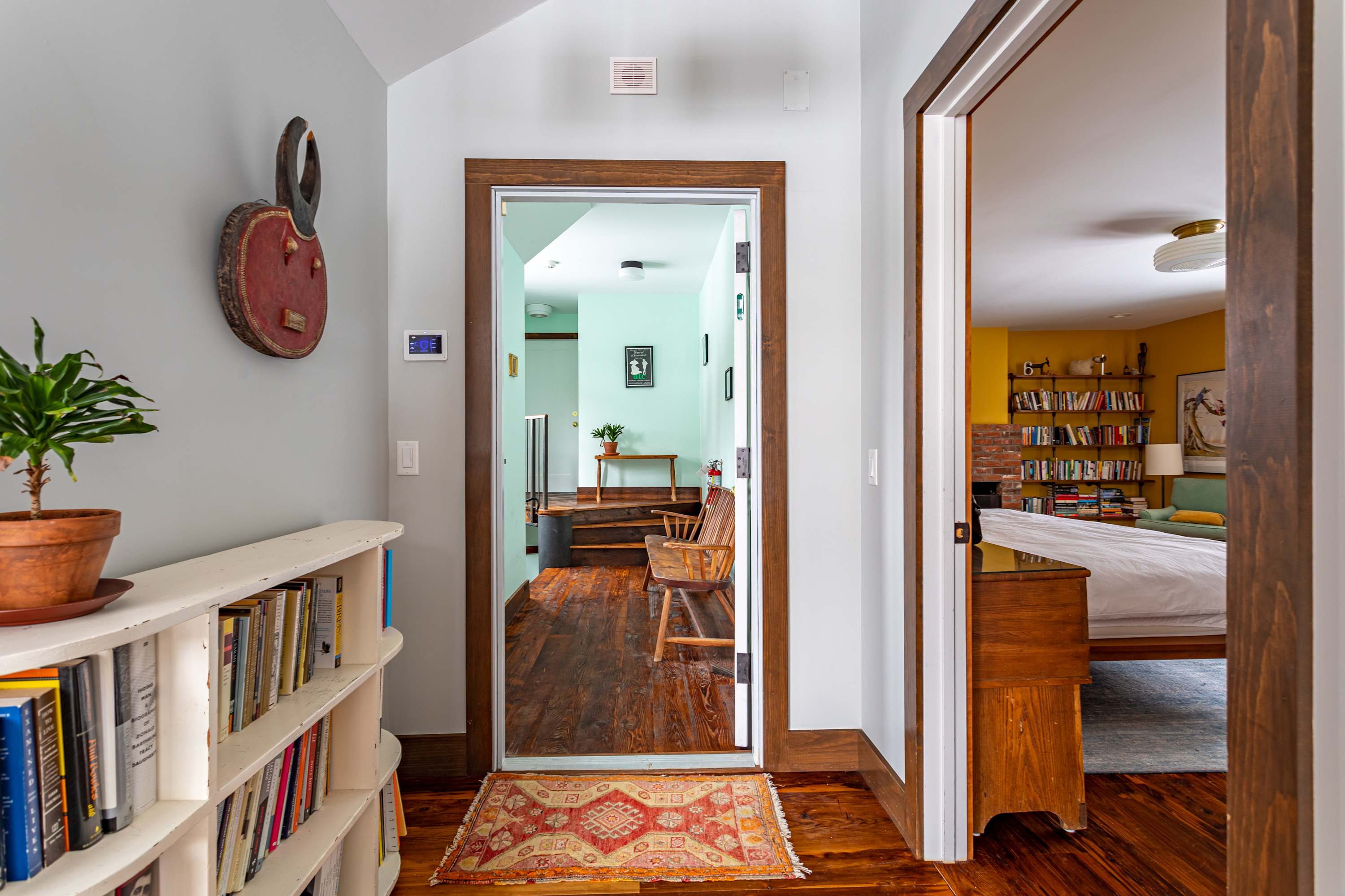 Bright hallway with light blue walls and dark hardwood floors, featuring a white bookshelf and a decorative wall hanging. Doorways lead to other rooms, including a yellow-walled bedroom.