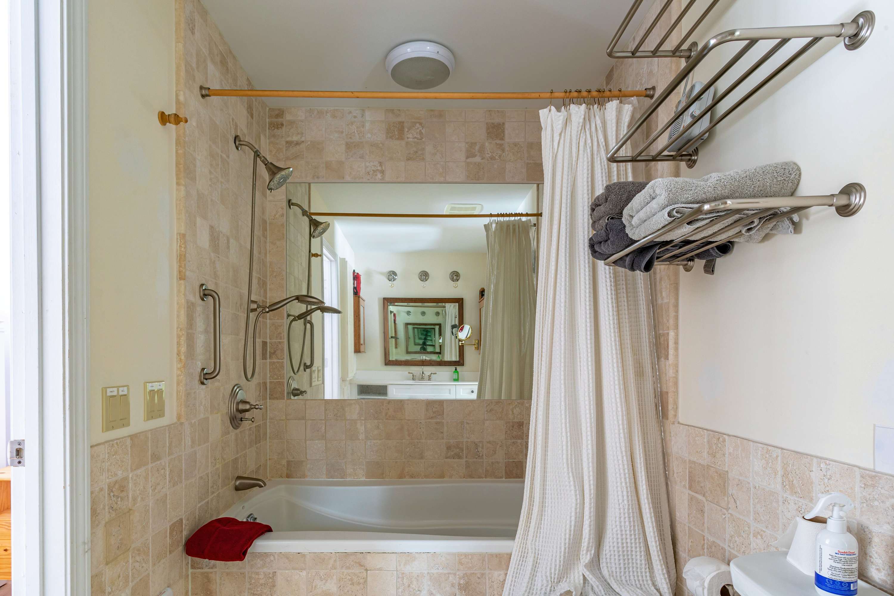 A bathroom with beige tiled walls features a white bathtub with a shower curtain, equipped with modern shower fixtures and a grab bar. A large mirror reflects the vanity area.