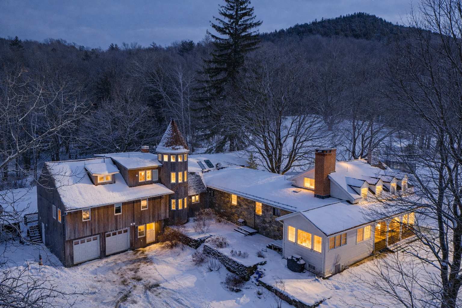 An aerial night view of the property covered in snow, with the house illuminated from within, showcasing its unique architecture including a round tower and multiple wings. The surrounding forest and mountains are also covered in snow.