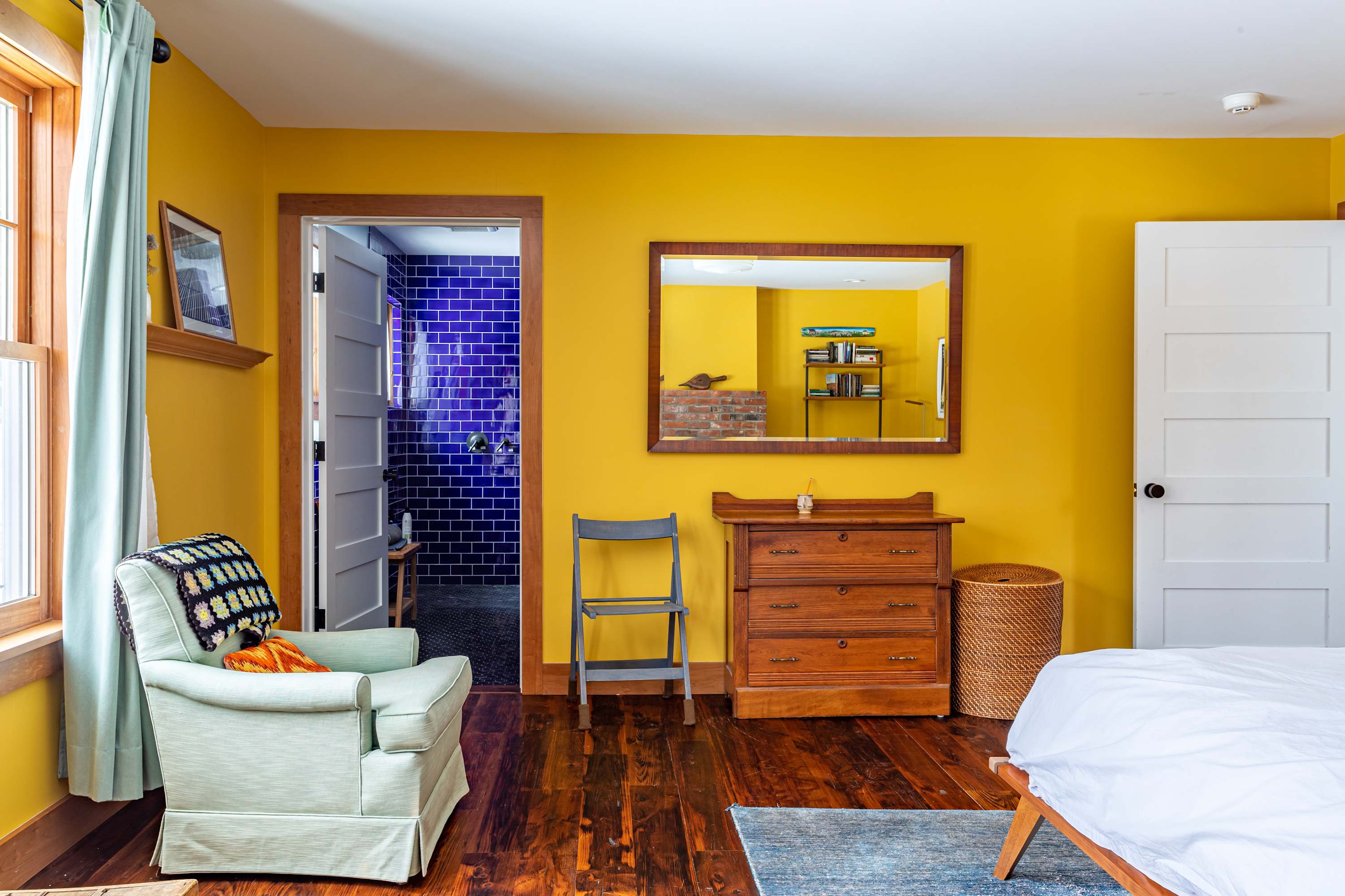 Vibrant yellow-walled bedroom with dark hardwood floors, featuring a light green armchair by a window and a wooden dresser. A doorway leads to a bathroom with striking blue tiles.