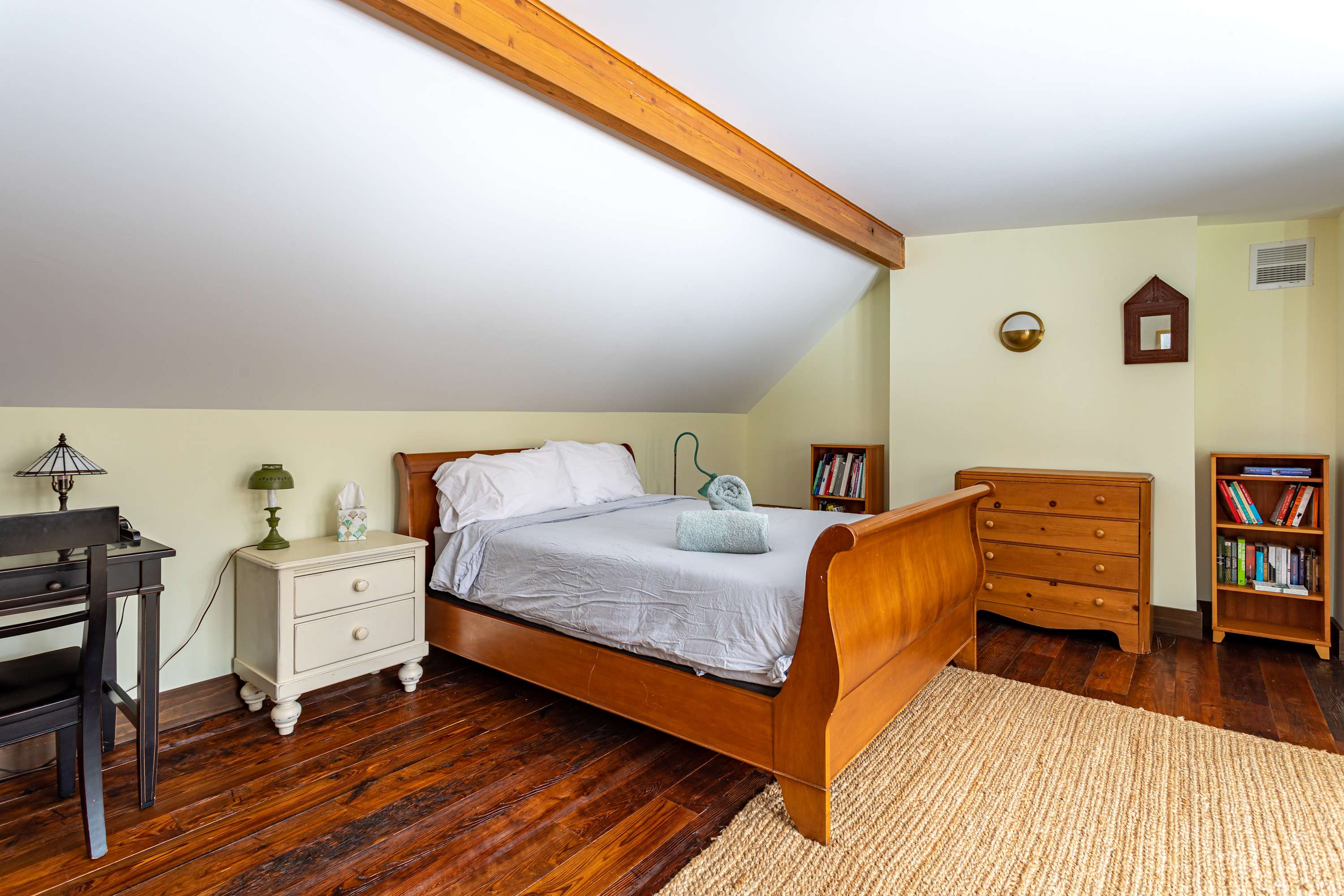 Cozy attic bedroom with a wooden sleigh bed, sloped white ceiling with exposed beam, dark hardwood floors, and light yellow walls. A small desk and nightstand are visible.