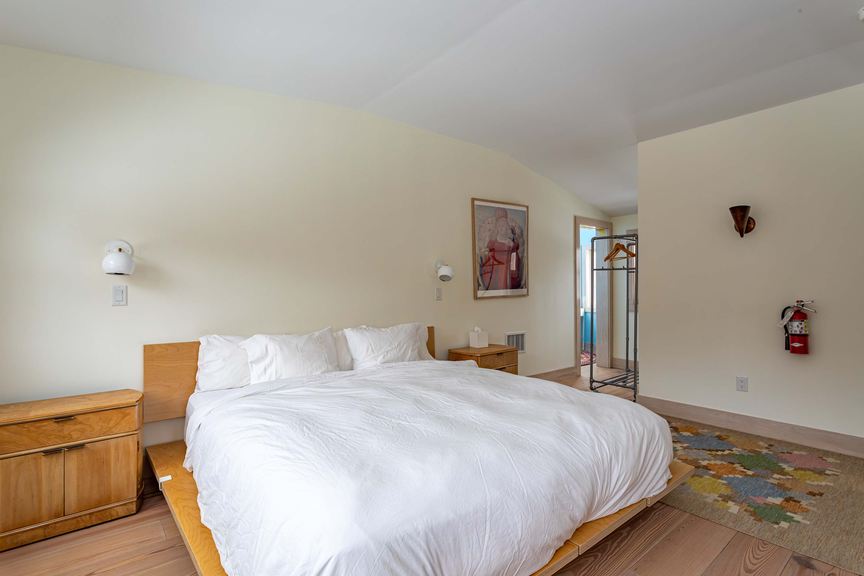 A wide shot of a bright bedroom with cream walls and hardwood floors, showcasing a modern wooden bed with white linens, two wooden nightstands, and a colorful rug. A clothing rack is visible in the corner.