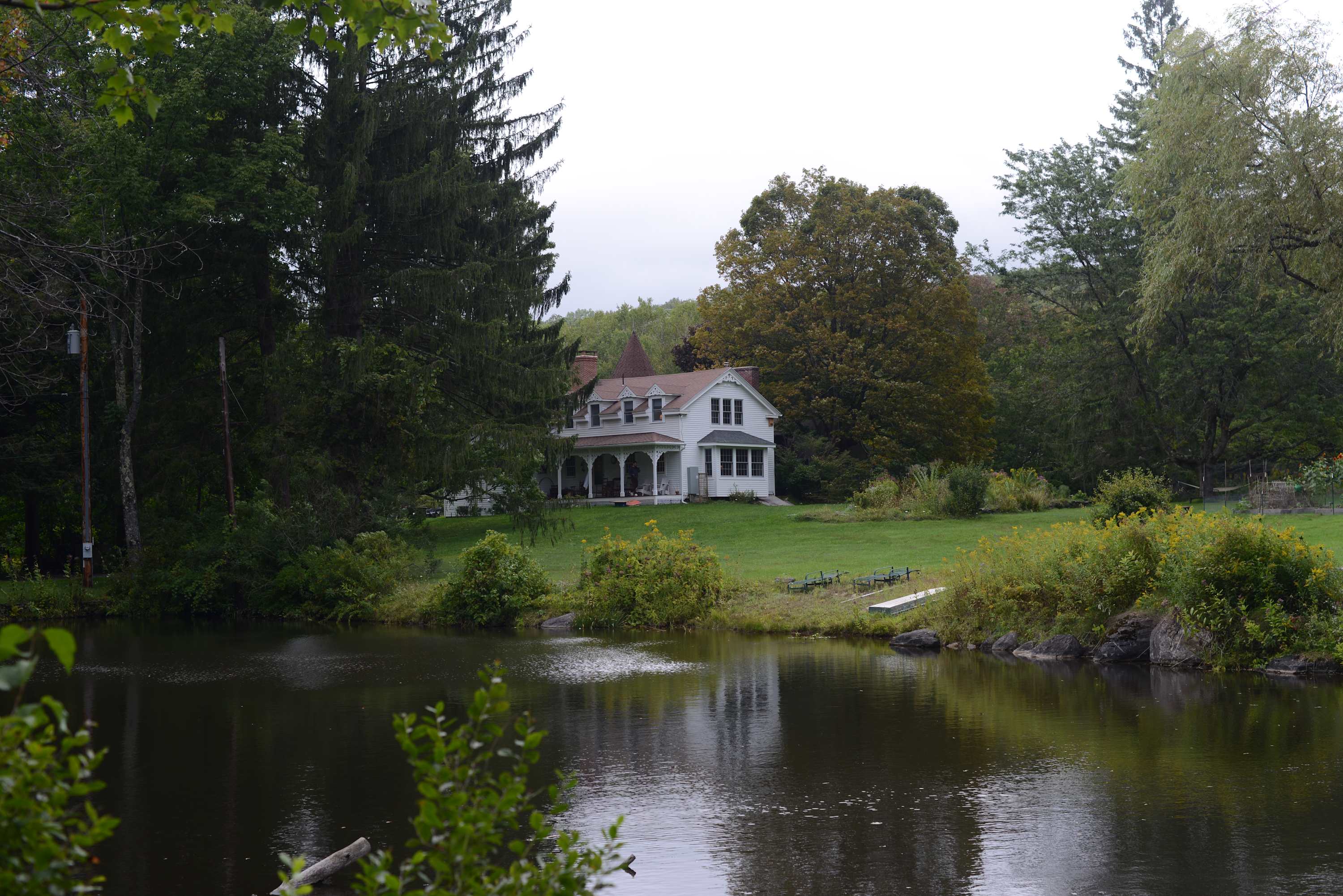 A scenic view of the white house with a distinctive turret, nestled among trees on a green lawn, reflected in the calm waters of a pond in the foreground.