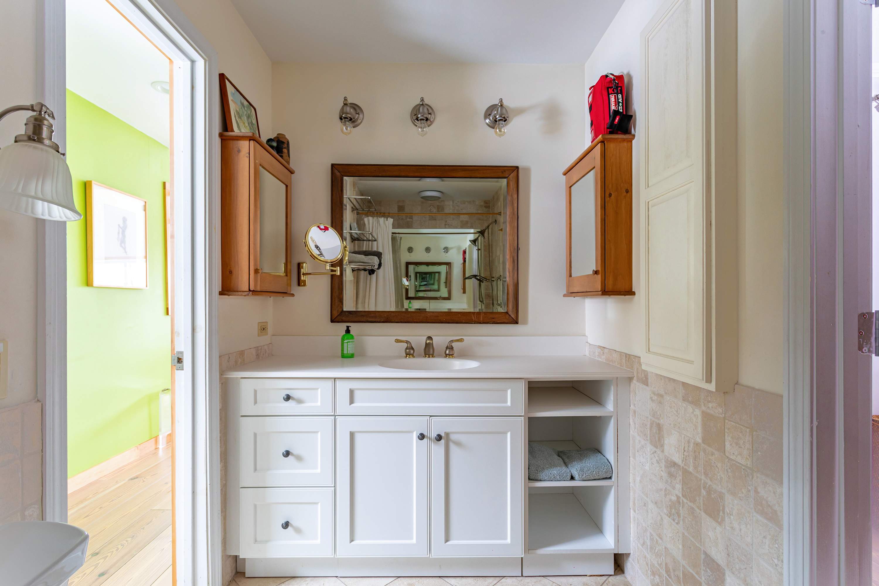 A bright bathroom vanity features a white countertop with an integrated sink, white cabinets, and open shelving, flanked by wooden medicine cabinets. A large mirror with sconce lighting hangs above.
