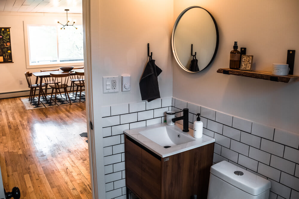 Bathroom vanity area with a modern wooden cabinet, white sink, black faucet, and a round black-framed mirror, set against white subway tile walls, with a view into the dining area.