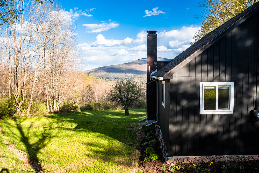Side view of a dark-colored cabin with white-trimmed windows and a prominent chimney, set against a backdrop of rolling green hills and distant mountains under a bright sky. The house is surrounded by a well-maintained grassy lawn.