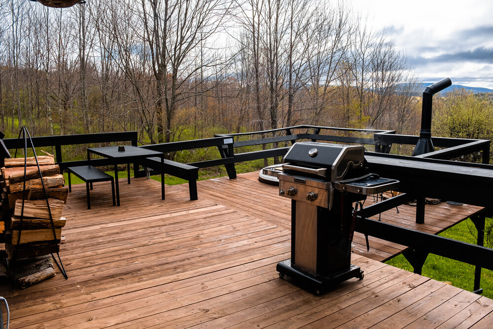 A spacious wooden deck features a gas grill, a small outdoor dining set, and a stack of firewood, all set against a backdrop of bare trees and distant hills under a cloudy sky.