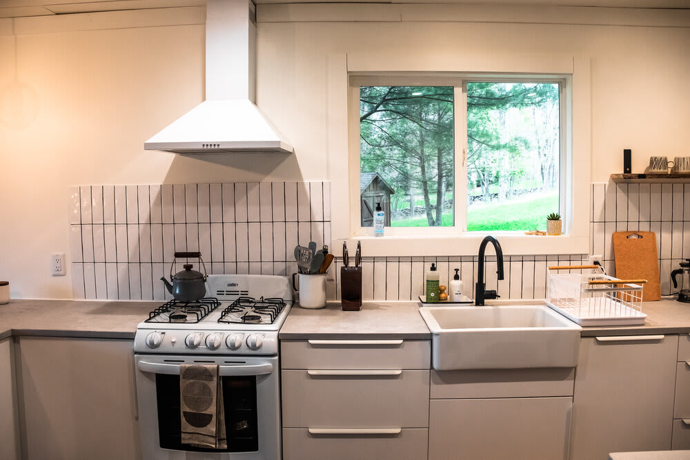 Close-up of the kitchen counter area featuring a white gas range, a white farmhouse sink with a black faucet, and white subway tile backsplash with dark grout, under a large window.