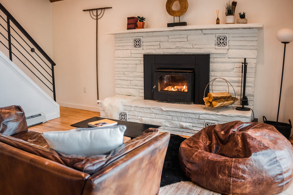 Cozy living room featuring a white stacked stone fireplace with a wood-burning insert, flanked by a brown leather sofa and a large brown bean bag chair, all resting on a patterned area rug.