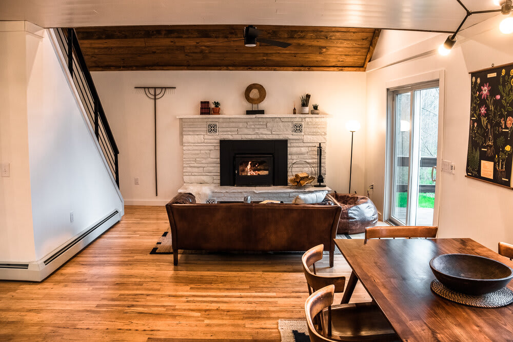 Wide shot of the open-concept living area with a white stacked stone fireplace, a brown leather sofa, and a dining area with a wooden table and chairs, all under a vaulted wooden ceiling.