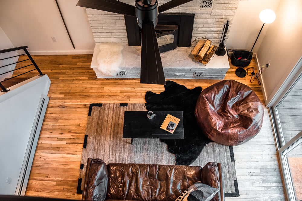 Overhead view of a cozy living room featuring a brown leather sofa, a large brown bean bag chair, and a black coffee table on layered rugs. A white stone fireplace and a prominent black ceiling fan complete the rustic-modern aesthetic.