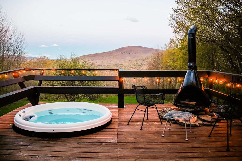 A wooden deck at dusk features a bubbling hot tub and a black outdoor fireplace, illuminated by string lights. The scene offers a serene view of a mountain landscape.