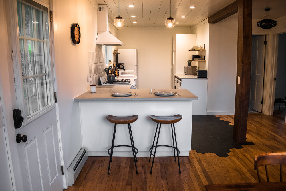 View of the kitchen bar with two wooden bar stools, separating the kitchen from the dining area, with a white exterior door visible on the left and a refrigerator in the background.