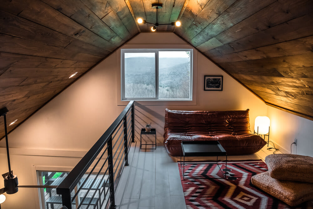 Loft lounge area with a low-profile brown leather sofa, small black side tables, and a large window offering panoramic views of a forested mountain landscape. The space features a sloped wooden plank ceiling and a black metal railing.