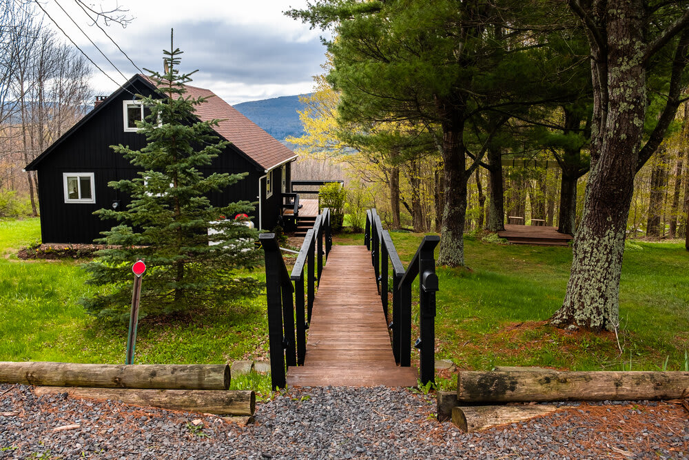 A wooden bridge with black railings leads towards the dark-colored cabin, nestled among tall trees and a vibrant green lawn. The entrance path is covered in gravel and wood logs, creating a rustic approach.