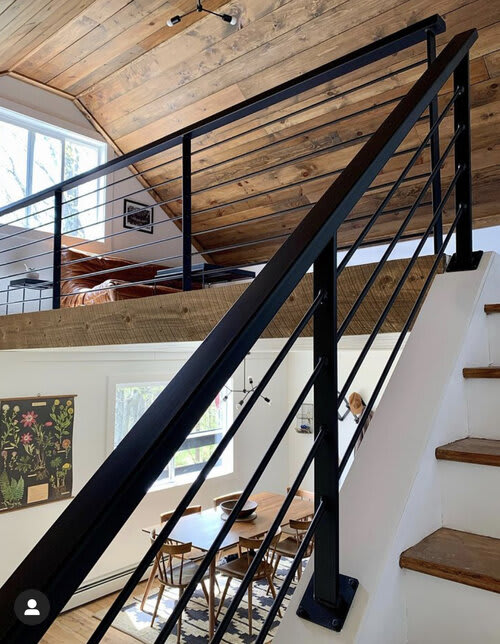 Elevated view from a loft or staircase, showcasing the open-concept living and dining areas below with black metal railings, hardwood floors, and a vaulted wooden ceiling.