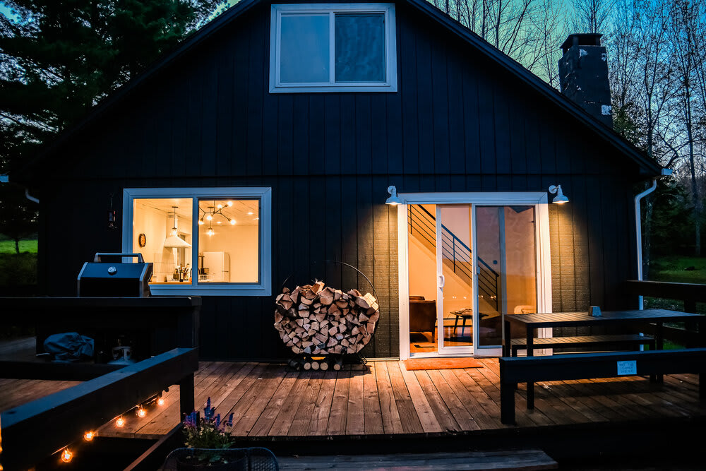 The back exterior of a dark-colored cabin at dusk, featuring a large wooden deck with a grill and stacked firewood. Illuminated sliding glass doors and windows reveal the interior.