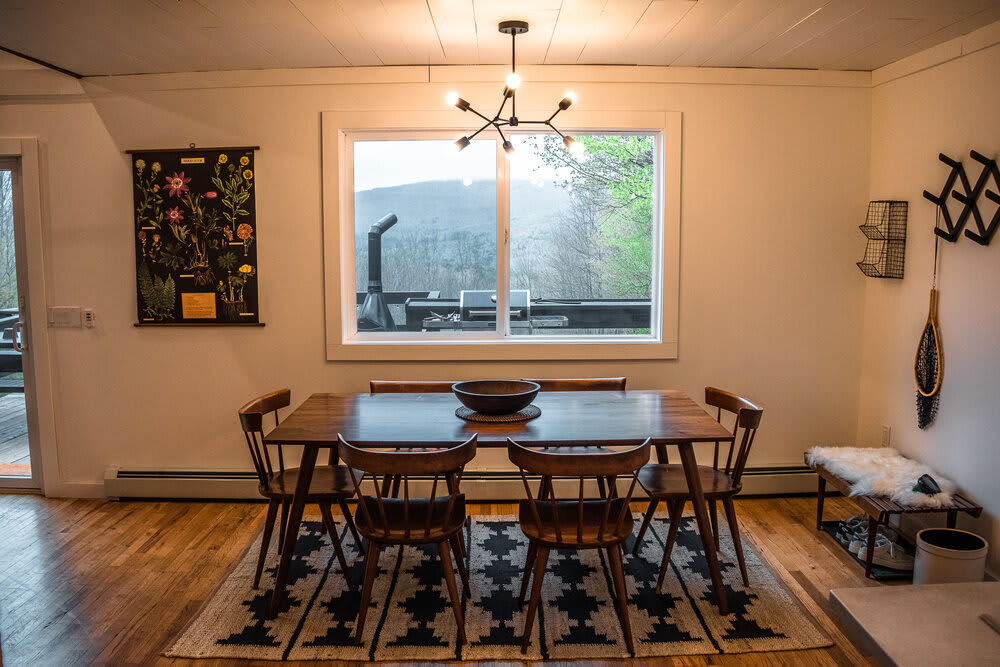 Dining area featuring a dark wooden rectangular table with six matching wooden chairs, centered on a black and white geometric rug, with a large window overlooking a lush green landscape.