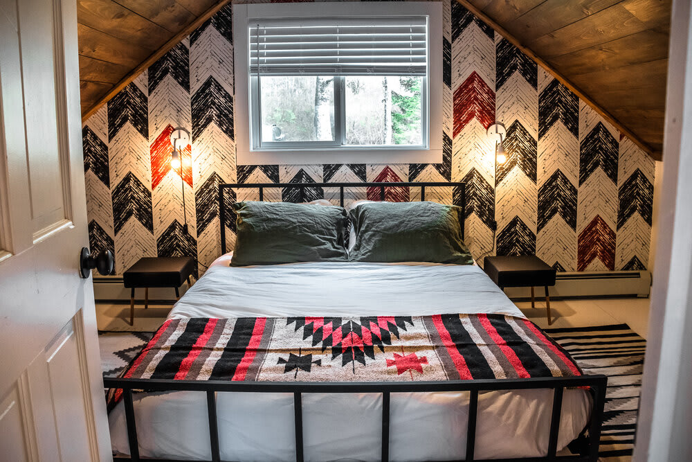 Inviting bedroom featuring a black metal bed frame with green pillows and a patterned blanket. The room is characterized by bold chevron-patterned wallpaper in red, black, and white, flanked by two small black nightstands and wall-mounted lights.