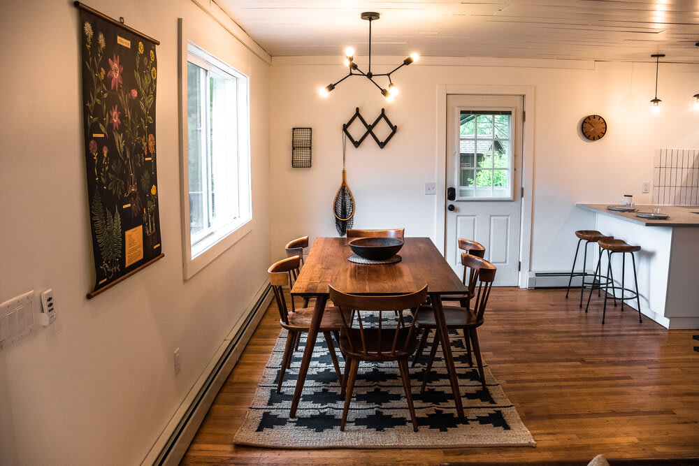 Dining area with a dark wooden table and chairs on a patterned rug, adjacent to a white exterior door and a view into the kitchen bar area, showcasing the open layout.