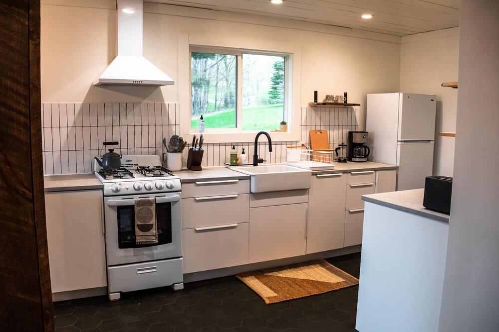 Modern kitchen with white flat-panel cabinets, light gray countertops, a white gas range with a matching hood, and a large window above a farmhouse sink, all on dark hexagonal tile flooring.