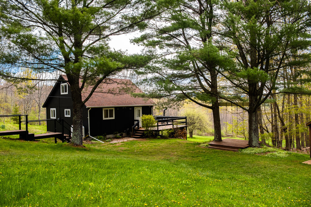 Expansive view of the dark-colored cabin with a visible deck, situated in a spacious green yard dotted with mature trees. The property is surrounded by natural foliage, offering a serene outdoor setting.