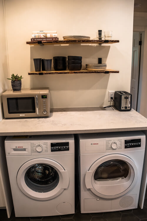 Dedicated laundry area equipped with two Bosch washer and dryer units beneath a light gray countertop. Floating wooden shelves above hold kitchenware, a microwave, and a toaster, indicating a combined utility and kitchenette space.