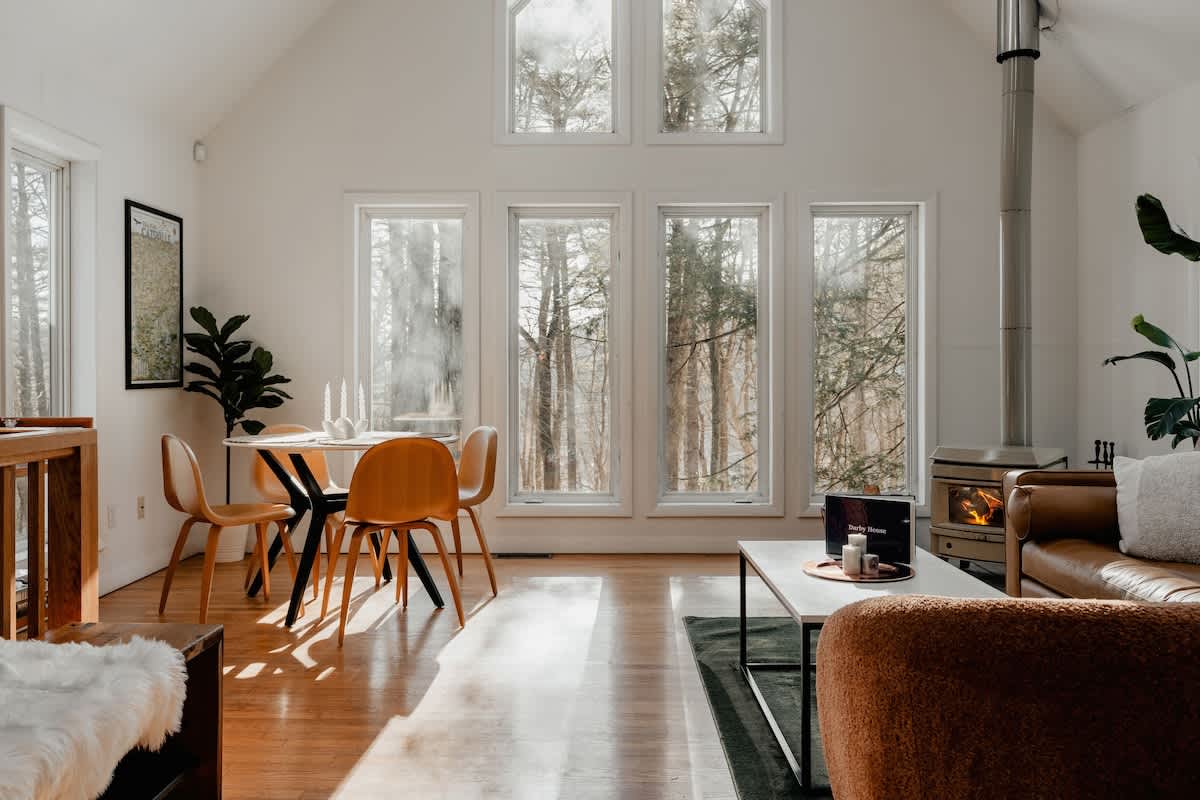 Bright living area with vaulted white ceilings and large windows offering forest views. A modern dining table with four wooden chairs is to the left, and a cozy seating area with a leather couch and wood-burning stove is to the right.