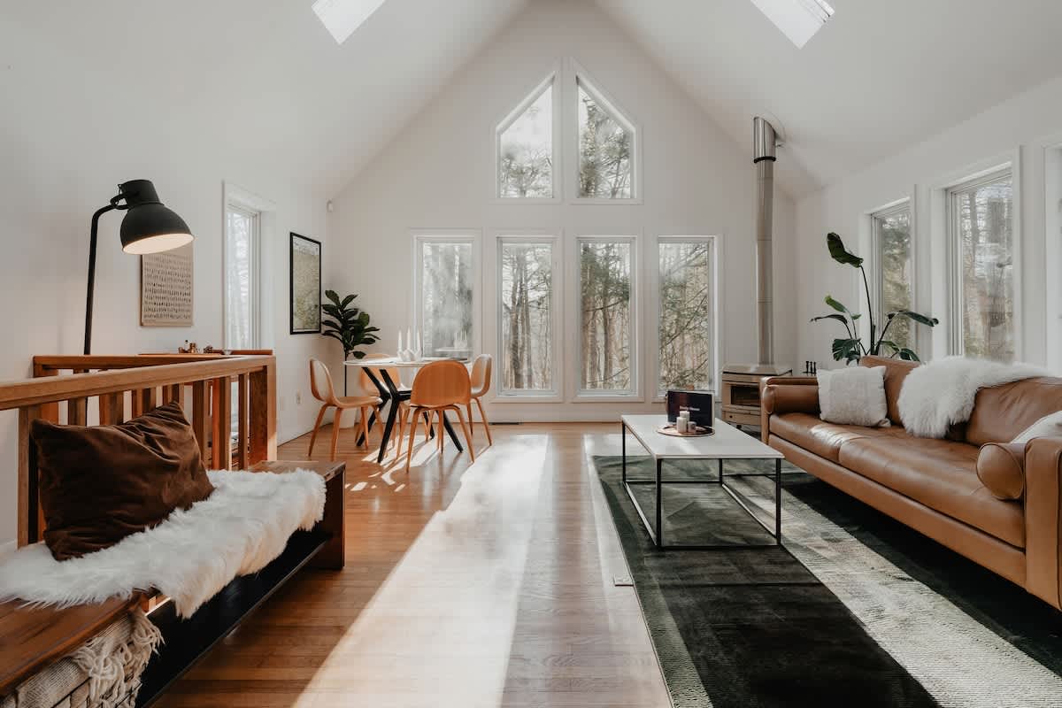 Sunny living and dining area with hardwood floors, a brown leather sofa, and a dining table with wooden chairs. Large windows offer views of the surrounding trees, and a wood-burning stove is visible.