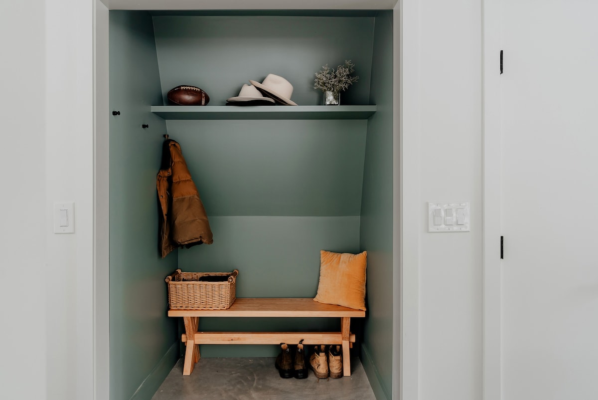 A cozy mudroom nook with light green walls features a wooden bench, coat hooks with a brown jacket, and a shelf displaying hats, a football, and a small plant. A woven basket and boots are placed below the bench.