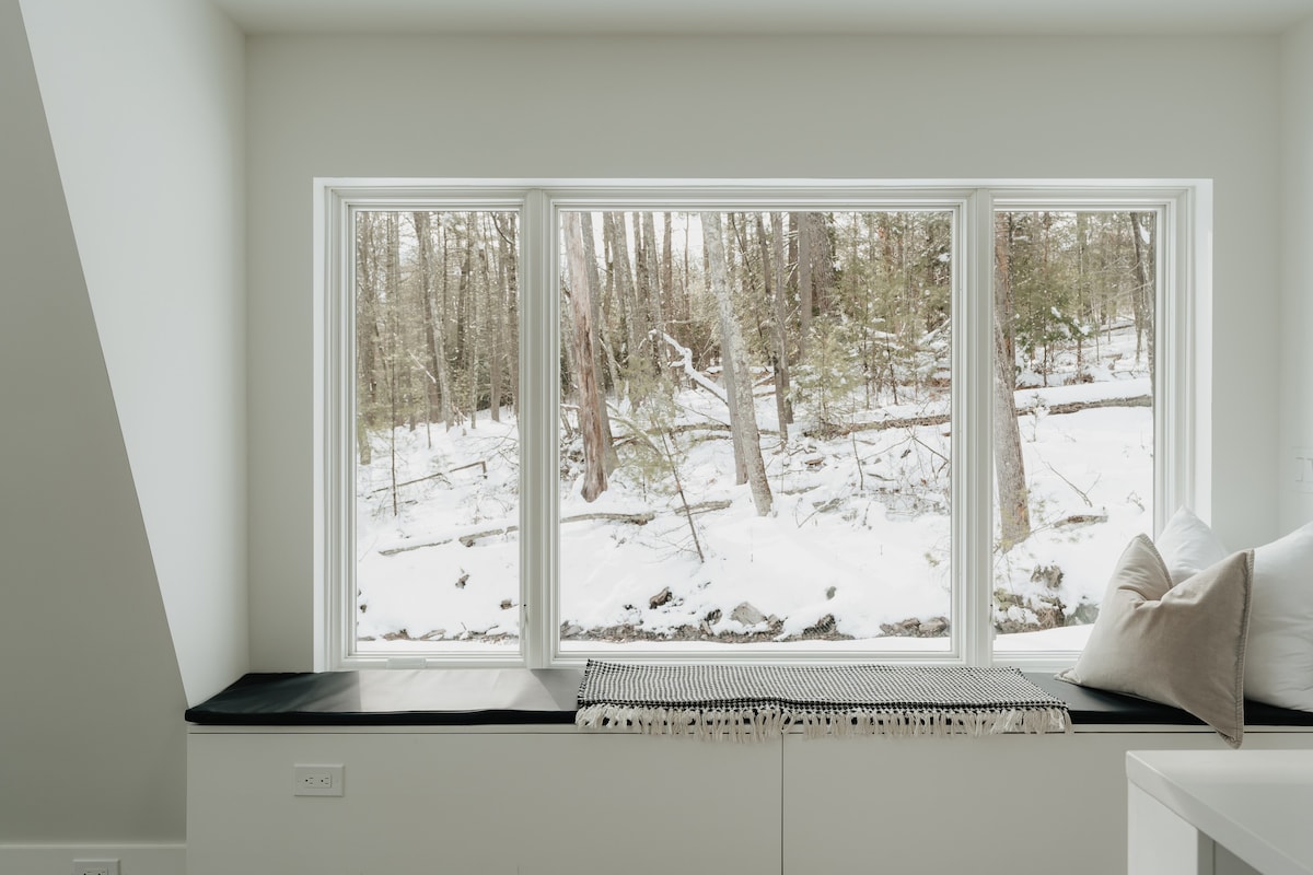 A cozy window seat with a black cushion and pillows offers a serene view of a snow-covered forest through large horizontal windows. A patterned throw blanket adds texture to the seating area.