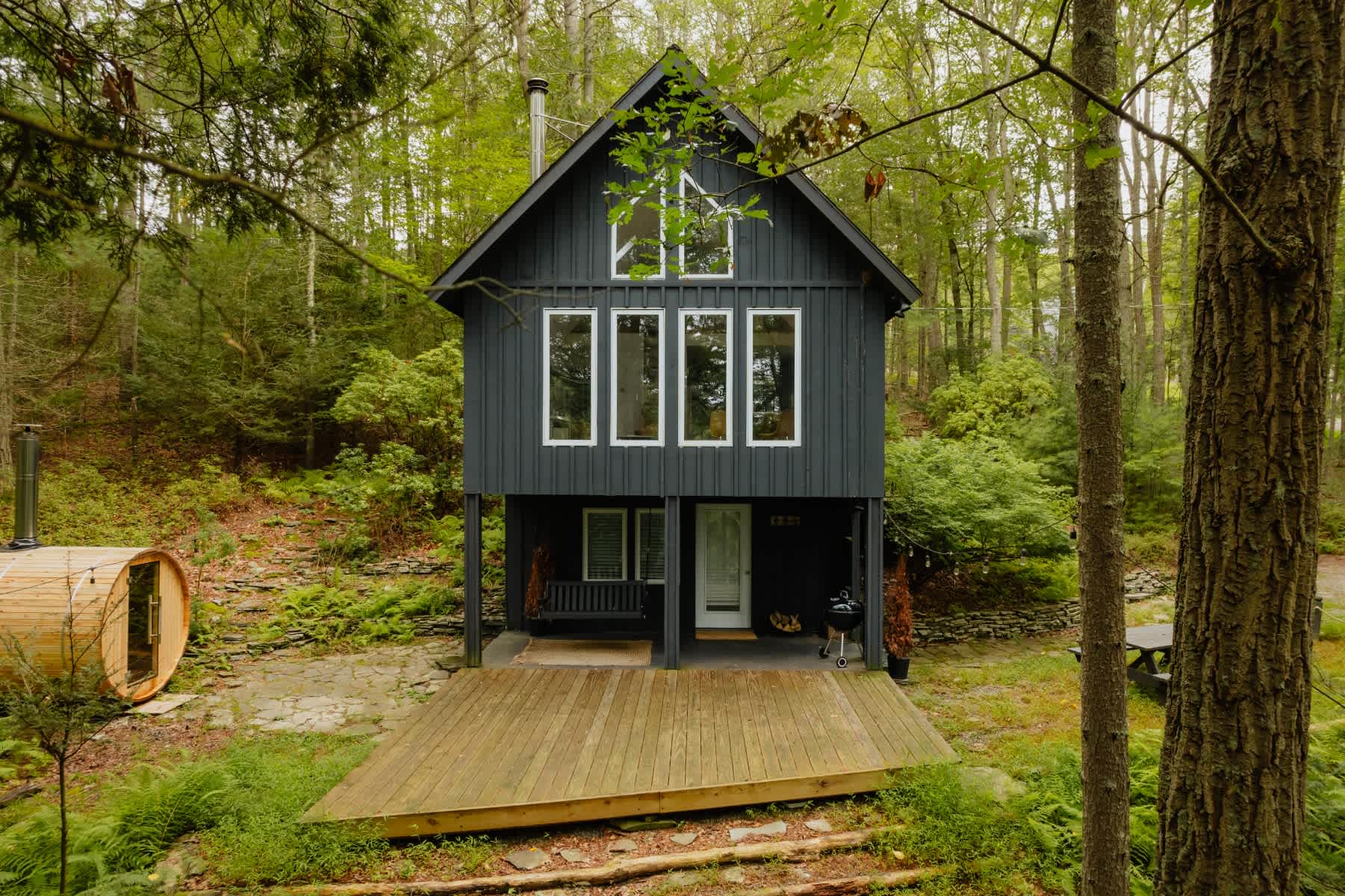 Dark grey cabin with multiple windows and a wooden deck, nestled in a lush green forest with ferns and tall trees. A barrel sauna is visible to the left, and a porch swing is under the deck.