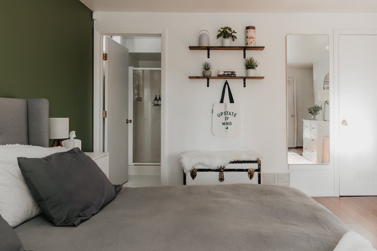 Another view of the bedroom, showing white walls with two wooden floating shelves holding plants and books. A white trunk with a faux fur throw sits below, and a full-length mirror reflects the room. A doorway leads to an en-suite bathroom.