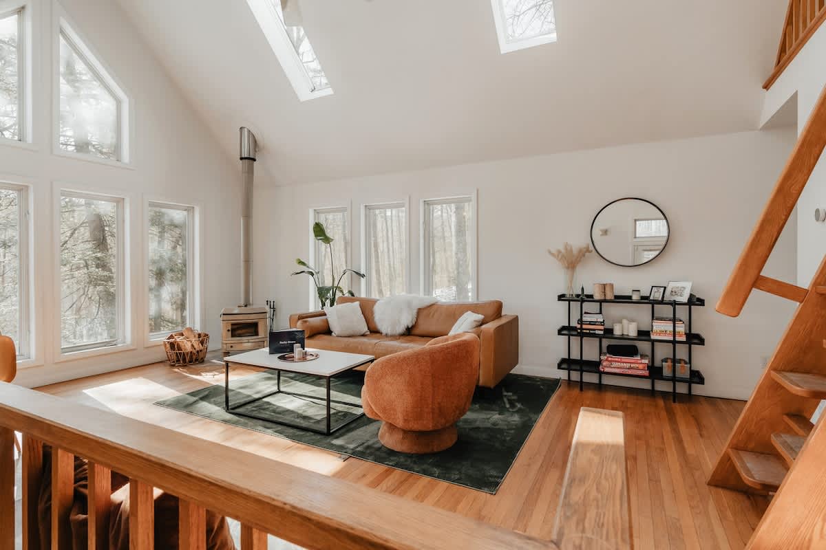 Inviting living room featuring a tan leather sofa, an orange swivel armchair, and a white coffee table on a dark green rug. Skylights and large windows illuminate the space, which also includes a black shelving unit and a round mirror.