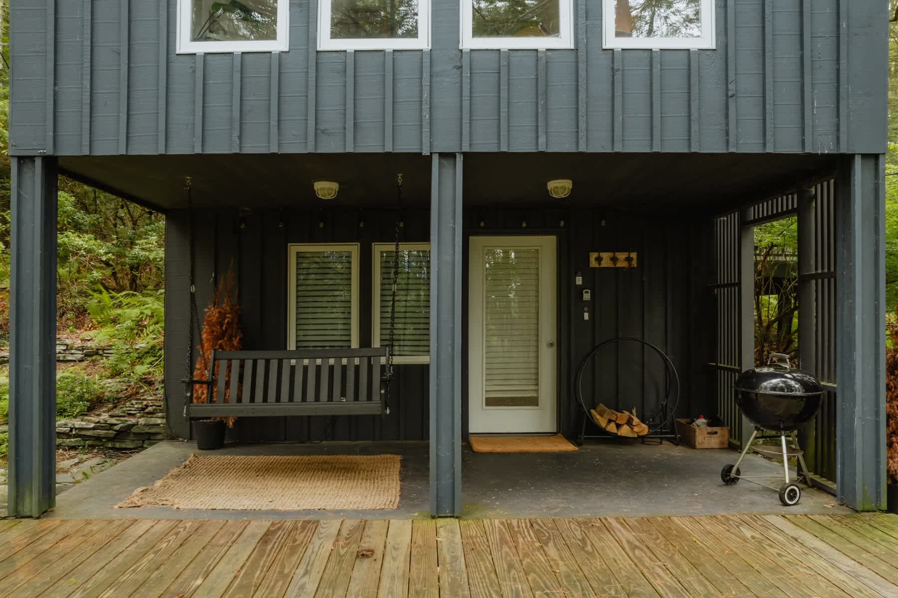 Covered outdoor porch area under the house, featuring a wooden swing, a white entry door, and a charcoal grill. The dark siding of the house and a wooden deck floor complete the rustic outdoor space.