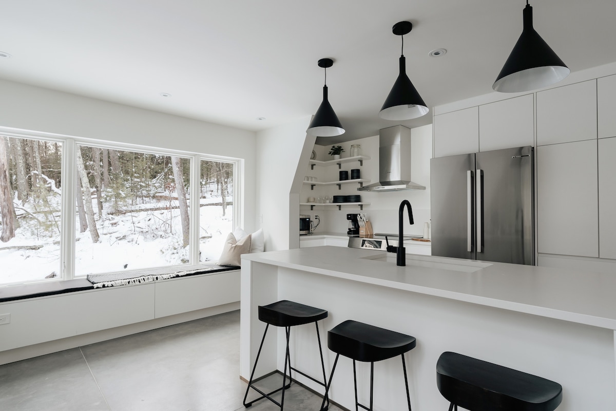 Modern kitchen with white flat-panel cabinetry, stainless steel appliances, and a large island with a black sink and three black bar stools. Large windows offer a snowy forest view, and open shelving displays dishes.