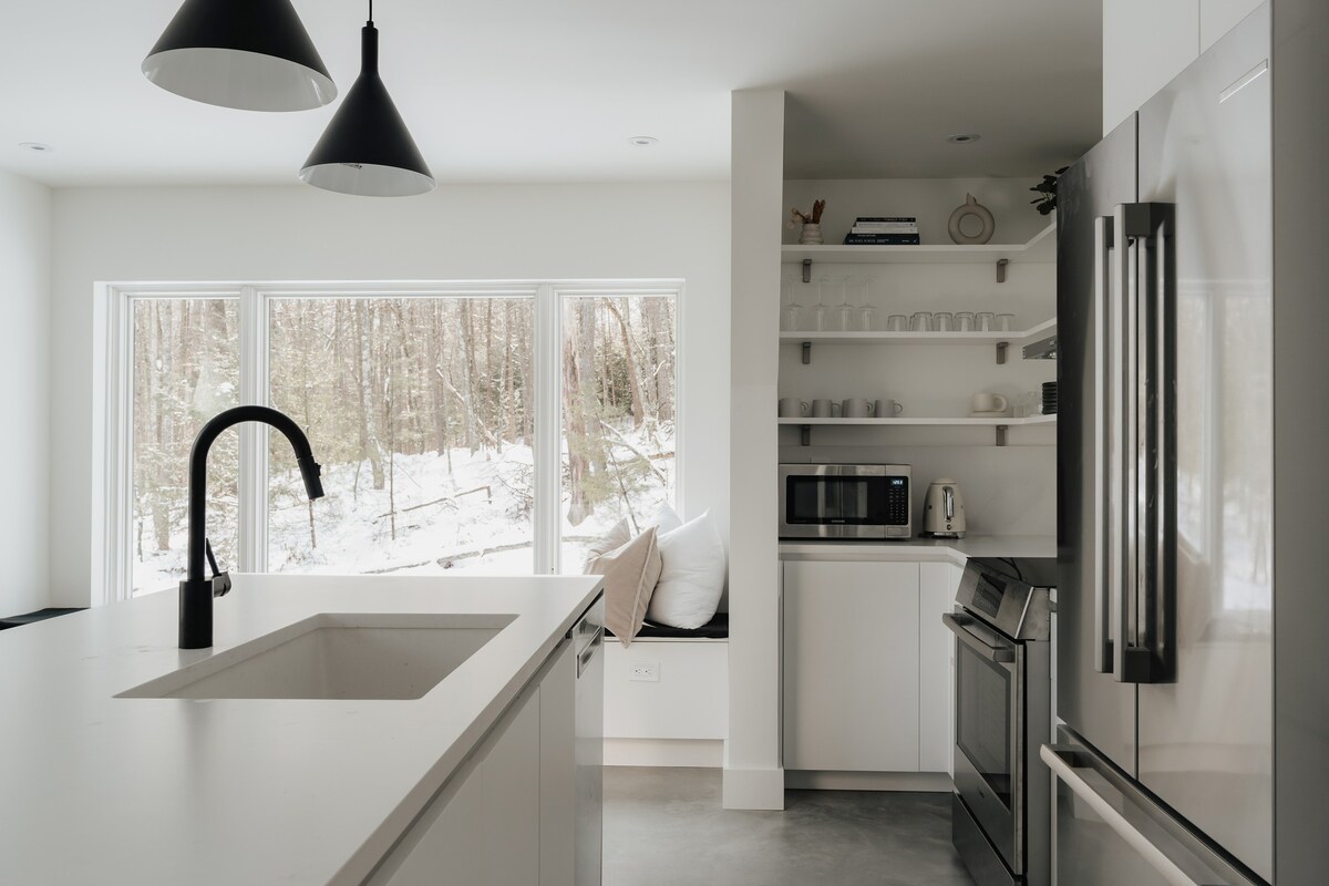 View of the kitchen island with a black gooseneck faucet and integrated sink, set against white countertops. In the background, large windows frame a snowy forest, with a window seat visible below.