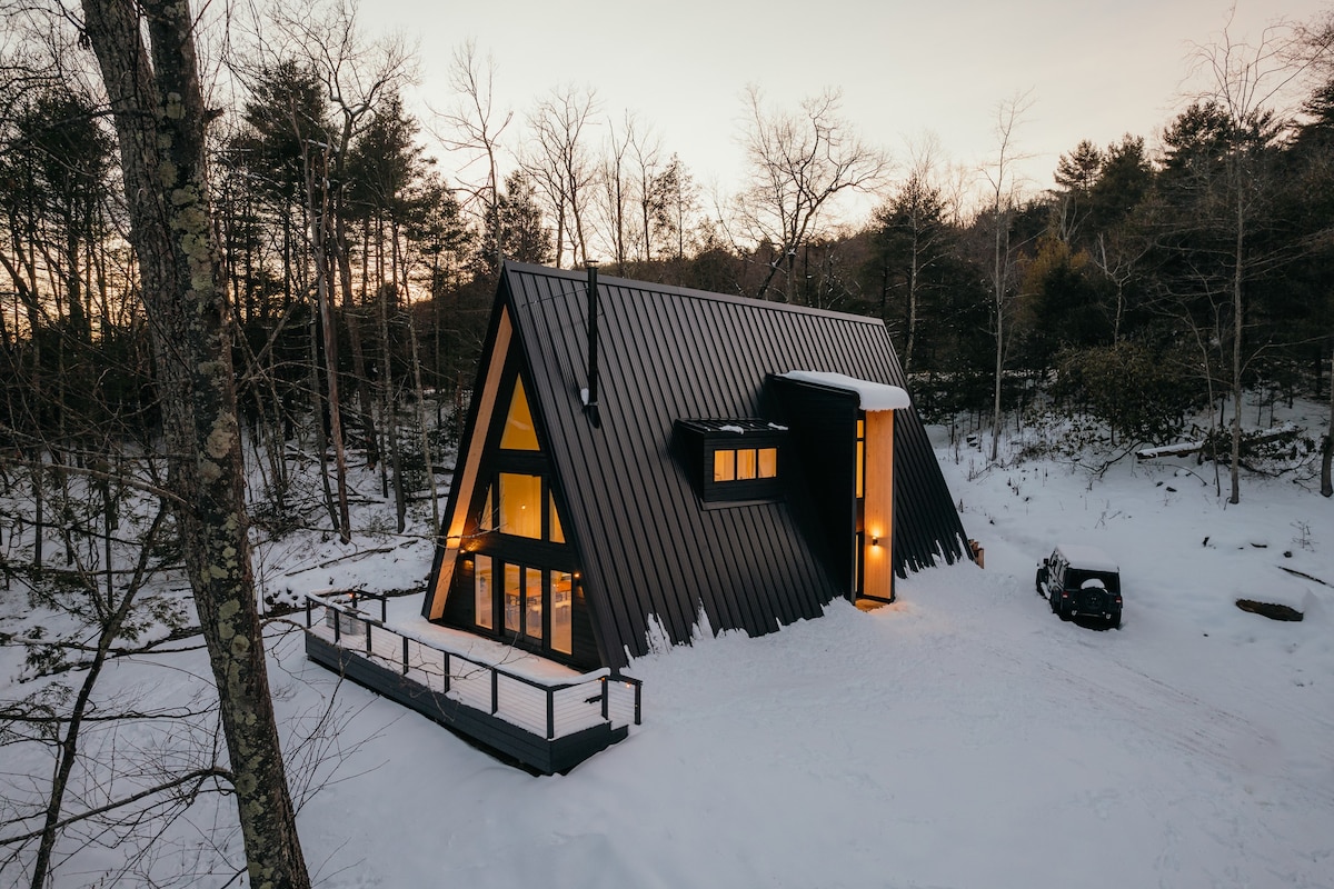 Winter aerial view of the black A-frame cabin, illuminated at dusk, surrounded by snow-covered trees and ground. The large windows glow warmly, and a vehicle is parked near the entrance.