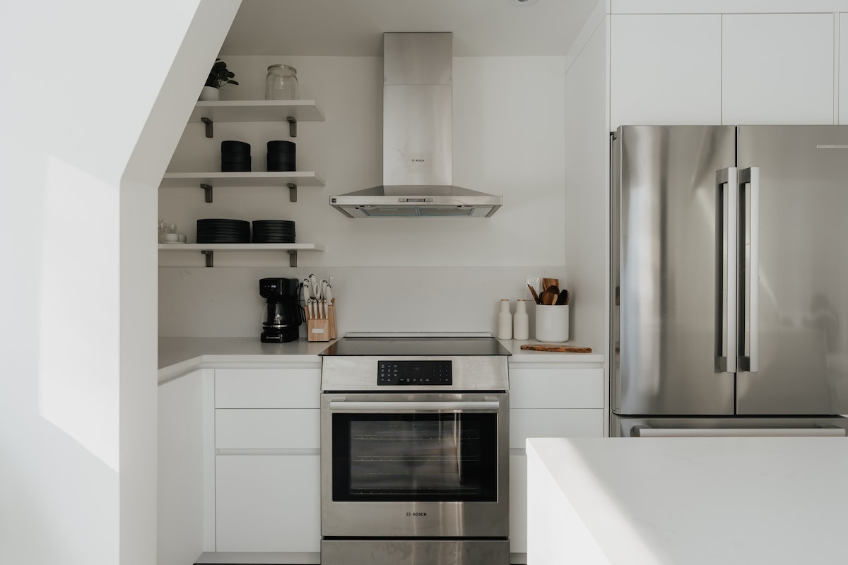 Detailed view of the kitchen showing stainless steel appliances including a refrigerator, oven, and range hood, integrated into white cabinetry. Open shelves display dishes and kitchen essentials, with a coffee maker on the counter.